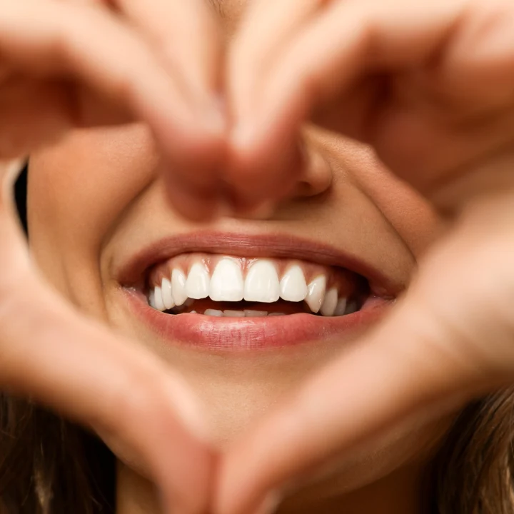 Mujer sonriente formando un corazón con las manos, mostrando dientes blancos y sanos.
