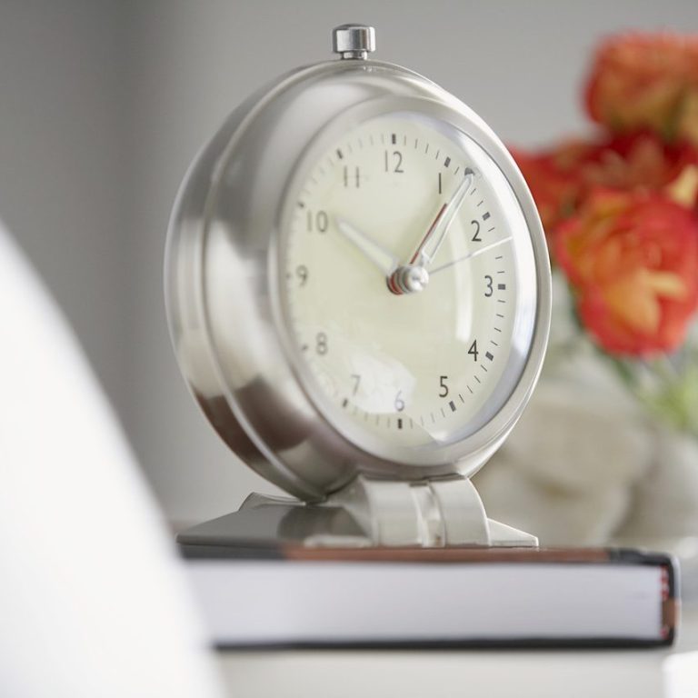 Reloj de mesa plateado junto a un libro y flores de colores.
