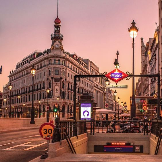 Salida de metro en una calle iluminada, con edificios y un atardecer de fondo.